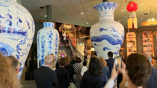La cérémonie d'ouverture de l'hôtel, avec des discours prononcés depuis les escaliers du hall, est en cours. De gigantesques répliques de vases chinois font partie de la création d'un environnement rappelant les Indes orientales.