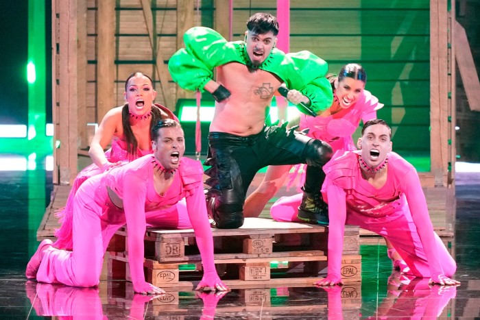 Un homme portant des manches bouffantes vertes chante entouré de personnes vêtues de tenues roses choquantes.