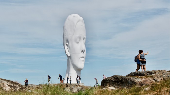 Une gigantesque sculpture blanche représentant une tête de femme aux yeux fermés. Au premier plan, des silhouettes humaines sont écrasées par l'échelle de la sculpture.